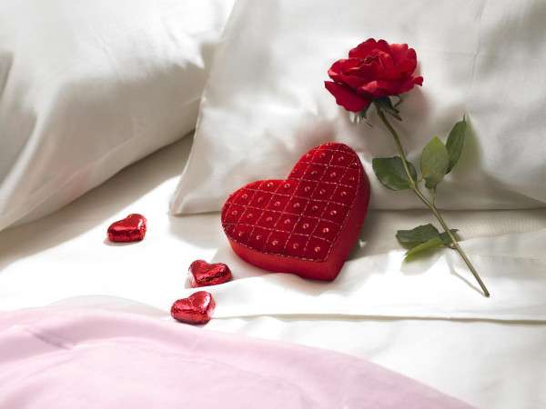 Valentine's Day romantic red rose and heart shaped candy box with red heart shaped chocolate candy on pillow of bed.